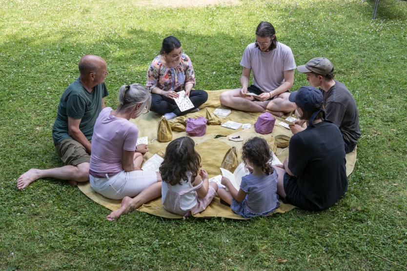 Eine Gruppe von Erwachsenen und Kindern sitzt im Kreis auf einer großen Decke im Gras und nimmt an einer gemeinsamen Aktivität teil.   A group of adults and children sits in a circle on a large blanket on the grass, taking part in a group activity. 