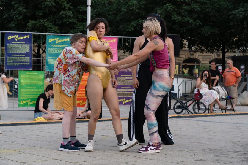 Eine Tanz-Performance im Freien, bei der sich mehrere Personen in unterschiedlicher Kleidung an den Händen halten und Bewegungen ausführen.  An outdoor dance performance where several performers wearing varied outfits interact and hold hands.