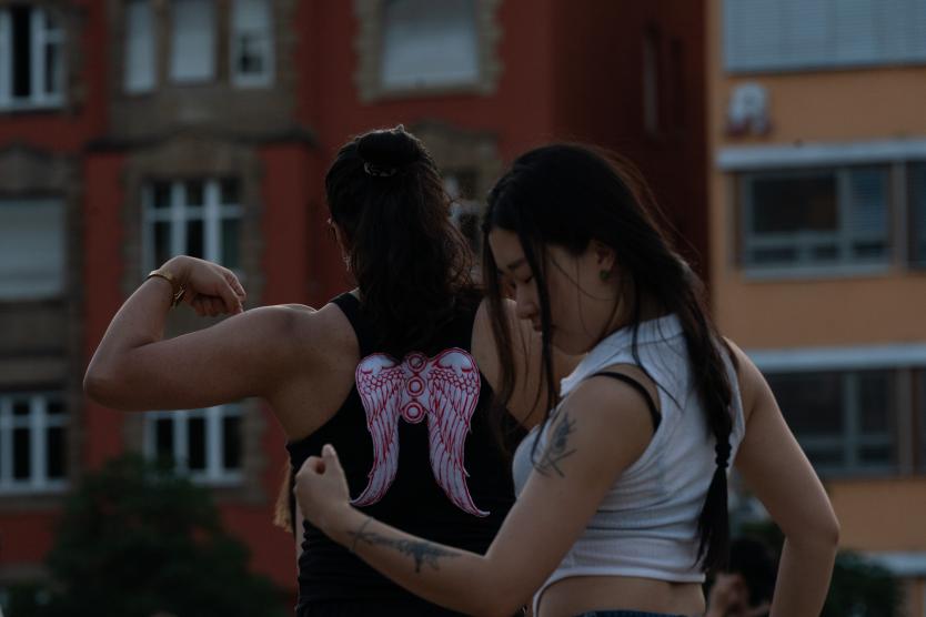 Nahaufnahme von zwei Personen von hinten im Abendlicht; eine Person spannt ihren Bizeps an, während die andere die Hand sanft auf ihren Rücken legt.  Close-up of two performers from behind at dusk, one flexing their bicep while the other gently places a hand on their back.