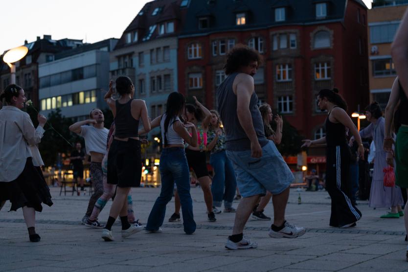 Eine Gruppe von Menschen tanzt am späten Abend auf einem gepflasterten städtischen Platz vor Wohngebäuden.   A group of people dancing outdoors on a paved public square during dusk, with residential buildings in the background. 