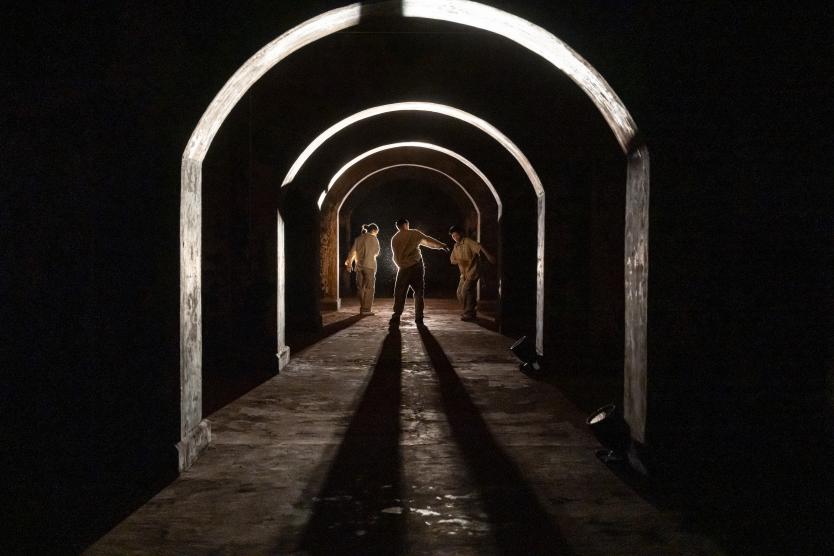 Drei Personen stehen in einem dunklen, tunnelartigen Raum mit mehreren hintereinander angeordneten Rundbögen. Eine Person in der Mitte streckt einen Arm zu einer anderen aus; starkes Gegenlicht wirft lange Schatten auf den Boden.      Three people stand in a dark, tunnel-like space with several rounded arches arranged one behind another. A person in the centre reaches out an arm towards another person; strong backlighting casts long shadows on the floor. 