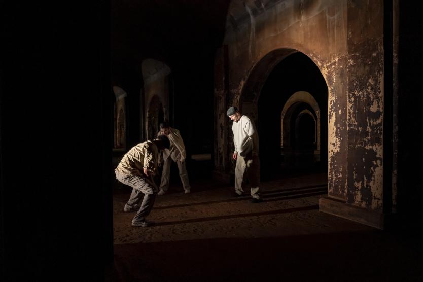 Drei Personen bewegen sich in einem dunklen Raum mit verwitterten Rundbögen und Säulen. Die Personen tragen helle, lockere Kleidung; warmes Seitenlicht erzeugt lange Schatten auf dem Boden.      Three people move through a dark room with weathered rounded arches and columns. The people wear light, loose-fitting clothing; warm side lighting creates long shadows on the floor. 