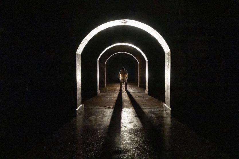 Eine Person in weißer Kleidung steht am Ende eines langen Ganges. Sie wird direkt von einem Scheinwerfer angestrahlt und die Silhouette wirft einen langen Schatten. A person dressed in white is standing at the end of a long hallway. A spotlight is shining directly on them, and their silhouette casts a long shadow.