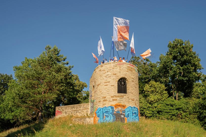 Ein gemauerter, historischer Aussichtsturm mit Graffiti auf einem grünen Hügel, auf dessen Spitze Personen große Fahnen im Wind halten.  A stone observation tower covered in graffiti on a grassy hill, with people at the top holding large flags in the wind.