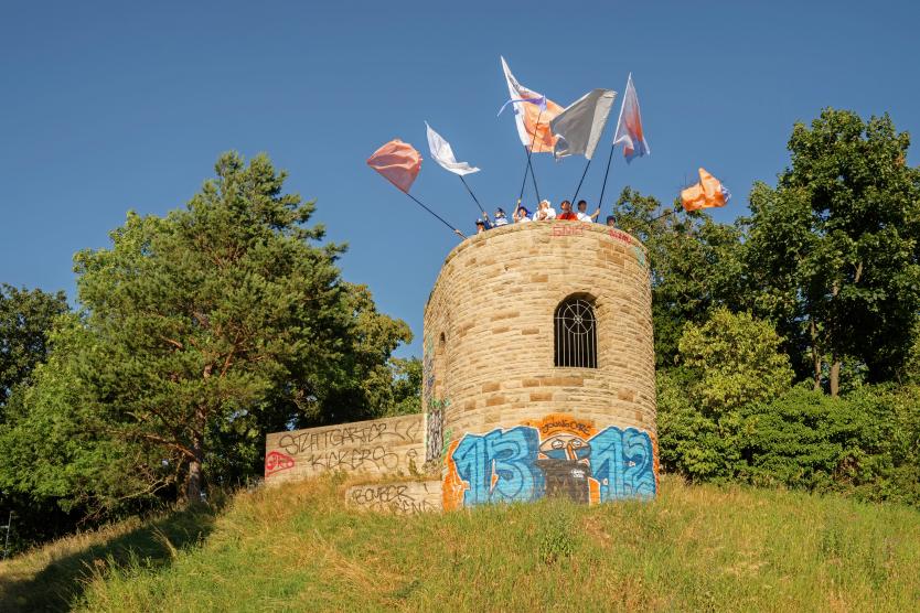 Eine Gruppe von Personen steht auf einem steinernen, mit Graffiti bemalten Aussichtsturm auf einem Hügel und hält farbige Fahnen in den blauen Himmel.   A group of people stands atop a stone observation tower adorned with graffiti on a grassy hill, holding colorful flags against a clear blue sky. 