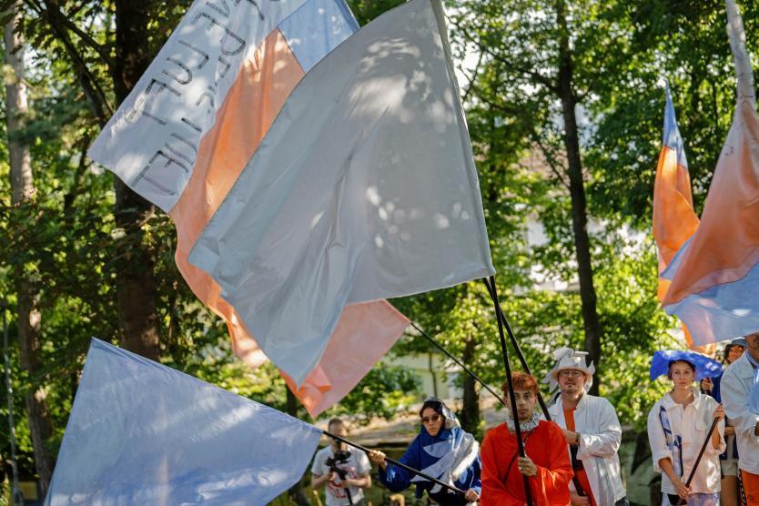 Nahaufnahme mehrerer Performer, die große hellblaue und orangefarbene Fahnen durch ein parkähnliches Gelände tragen.   Close-up of several performers carrying large light blue and orange flags through a park-like setting. 
