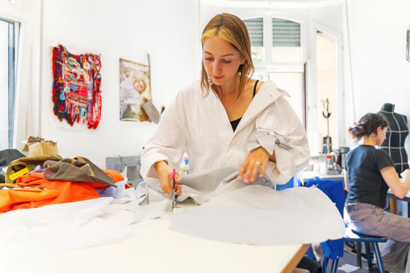 Eine Frau mit hellen Haarsträhnen arbeitet in einer Werkstatt an einem Tisch und schneidet hellem Stoff mit einer Schere zu.   A woman with highlighted hair works at a table in a workshop, cutting light-colored fabric with scissors. 