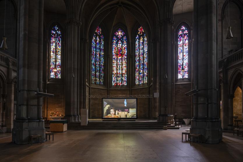 Eine Projektionsleinwand im Altarraum einer gotischen Kirche mit bunten Glasfenstern zeigt ein Video einer Person im Innenraum eines Autos.  A projection screen in the chancel of a gothic church with stained glass windows displays a video of a person inside a car.