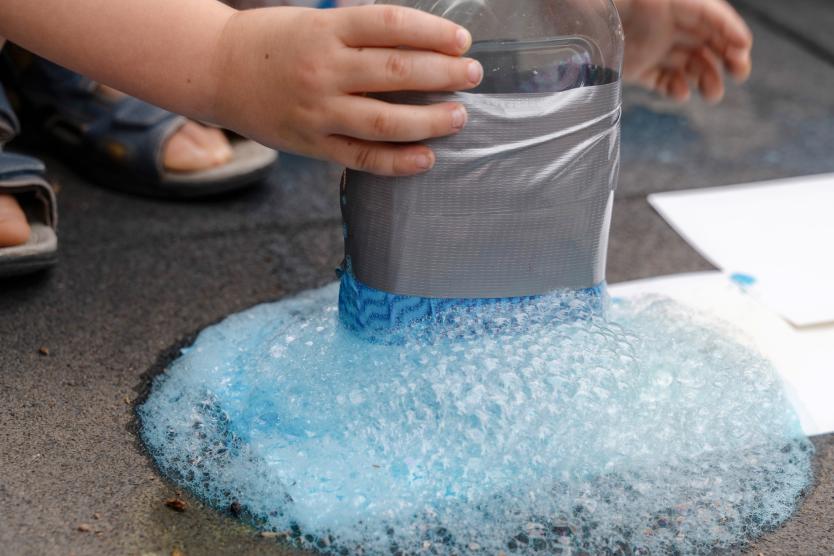 Nahaufnahme der Hände eines Kindes, das eine abgeklebte Plastikflasche hält, aus der hellblauer Seifenschaum auf den Boden quillt.  Close-up of a child's hands holding a taped plastic bottle creating a pool of light blue soap foam on the ground.