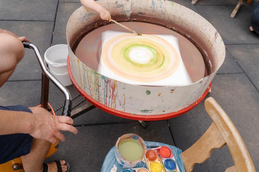 Draufsicht auf eine rotierende Töpferscheibe oder Drehscheibe, auf der ein Kind mit einem Pinsel farbige Kreise auf Papier malt.  Overhead view of a child using a paintbrush to create colorful concentric circles on paper spinning on a pottery wheel setup.