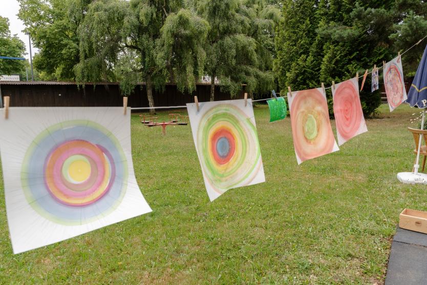 Auf einer Wäscheleine im Freien hängen mehrere Blätter Papier zum Trocknen, auf die bunte, kreisförmige Muster gemalt wurden.  Colorful circular paintings on paper hanging to dry on a clothesline outdoors in front of a green lawn.