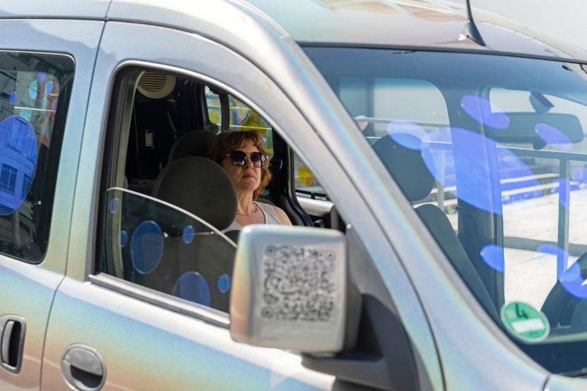 Eine Frau mit Sonnenbrille sitzt am Steuer eines Autos, dessen Fensterscheiben mit blauen, transparenten Kreisen dekoriert sind.  A woman wearing sunglasses seen through the window of a car decorated with semi-transparent blue circular stickers.