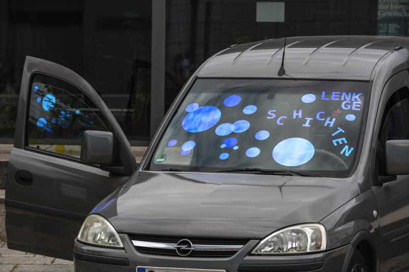 Nahaufnahme der Front eines grauen Autos, dessen Windschutzscheibe mit blauen Kreisen und dem Schriftzug „LENK GESCHICHTEN“ beklebt ist.Close-up of the front of a gray car with blue circles and the text "LENK GESCHICHTEN" applied to its windshield.