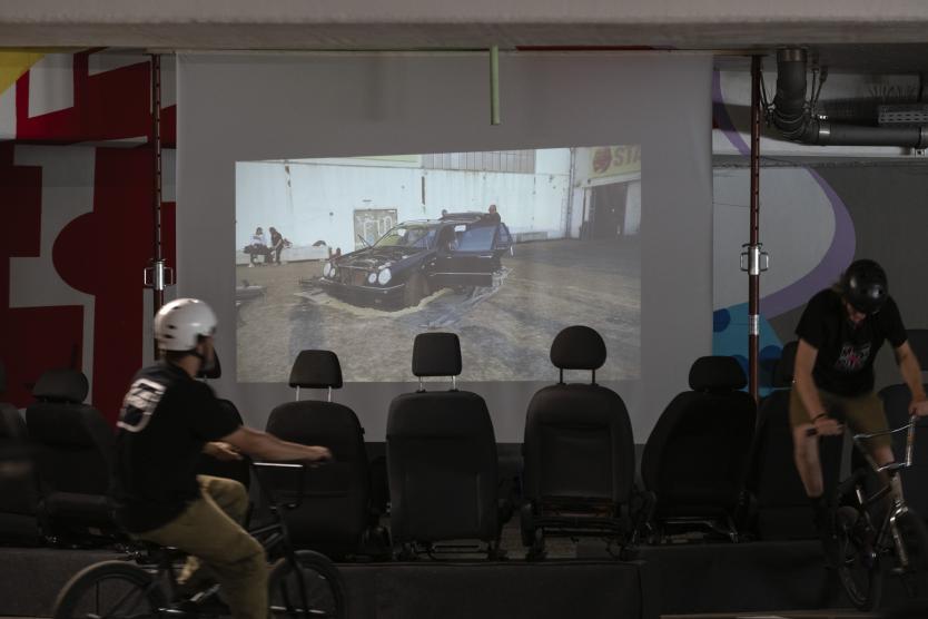Eine Videoprojektion an der Wand einer Indoor-BMX-Halle zeigt ein demontiertes Auto, während BMX-Fahrer im Vordergrund vorbeifahren.  A video projection on the wall of an indoor BMX facility shows a dismantled car, while BMX riders pass by in the foreground.