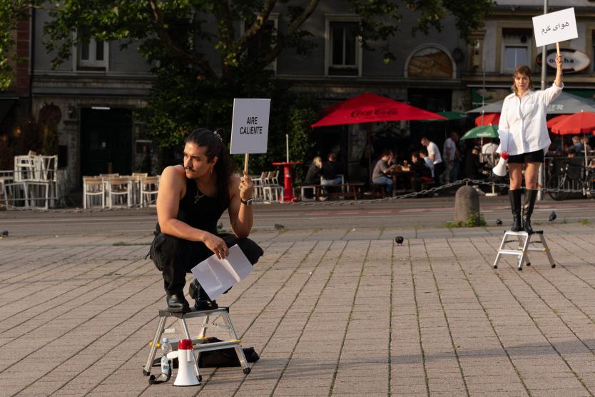 Auf einem gepflasterten Platz hocken zwei Performer jeweils auf kleinen Trittleitern: Im Vordergrund hockt ein Mann mit Schild „AIRE CALIENTE“ und ein Megafon zu seinen Füßen, im Hintergrund steht eine Person mit einem Schild in persischer Schrift.  On a paved square, two performers are positioned on small step ladders: in the foreground, a man crouches holding a sign reading "AIRE CALIENTE" with a megaphone at his feet, while another person stands in the background holding a sign in Persian script.