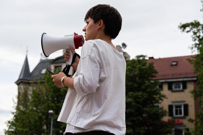 Seitenprofil einer Person mit kurzen dunklen Haaren und weißem Oberteil, die im Freien ein weiß-rotes Megafon an den Mund hält, um zu sprechen.  Side profile of a person with short dark hair and a white top, holding a white and red megaphone to their mouth outdoors.