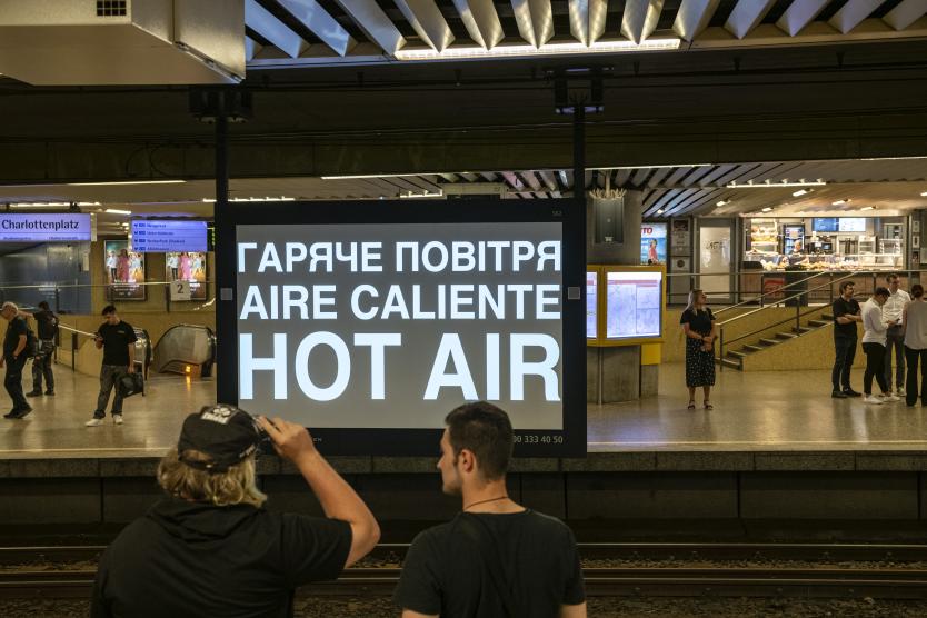 Ein großer digitaler Werbebildschirm an einem U-Bahnhof zeigt mehrsprachigen Text: „ГАРЯЧЕ ПОВІТРЯ / AIRE CALIENTE / HOT AIR“. Zwei Passanten im Vordergrund betrachten die Anzeige.  A large digital display board at a subway station displays multilingual text: "ГАРЯЧЕ ПОВІТРЯ / AIRE CALIENTE / HOT AIR". Two passersby in the foreground look at the screen.