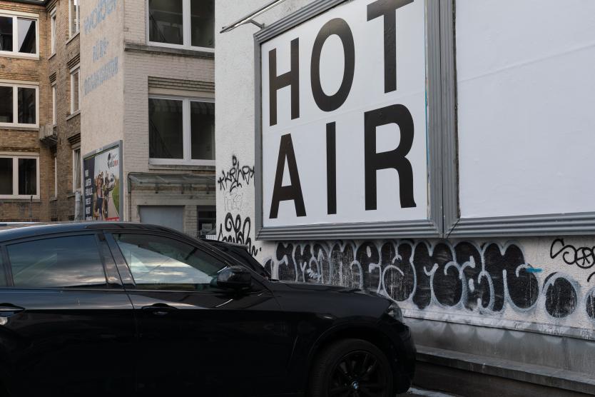 Ein Plakat mit der Aufschrift „HOT AIR“ in großen schwarzen Buchstaben hängt an der Fassade eines Gebäudes, vor dem ein schwarzes Auto geparkt ist.  A billboard reading "HOT AIR" in large black lettering is mounted on a building facade next to a parked black car.