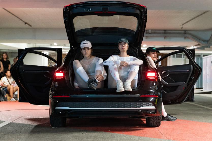 Zwei Performerinnen in weißer Kleidung sitzen im geöffneten Kofferraum eines schwarzen Autos in einem Parkhaus.  Two performers in white clothing sit inside the open trunk of a black car in a parking garage.