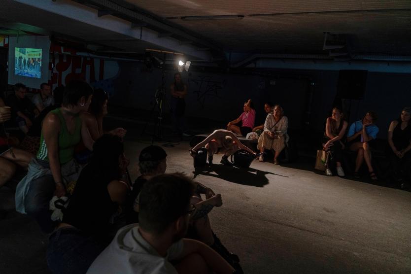 Ein Publikum sitzt im Halbdunkel einer Halle und beobachtet eine Performance-Künstlerin am Boden unter einem Scheinwerferspot.  An audience sits in the dim light of a hall, watching a performance artist on the floor illuminated by a spotlight.