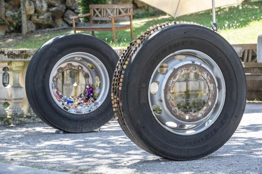 Zwei große Lkw-Reifen stehen im Freien auf einem Kiesweg, wobei einer mit Glitzerelementen verziert ist und der andere bunte Objekte im Felgeninneren zeigt.  Two large truck tires stand outdoors on a gravel path, one decorated with glittery elements and the other displaying colorful objects inside its rim.