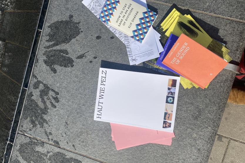 Auf einer Steinfläche liegen verschiedene Broschüren, Flyer und Bücher zu Themen wie Kunst und Feminismus, teils auf feuchtem Untergrund.   Various brochures, flyers, and books on art and feminism are arranged on a stone surface, partially wet. 