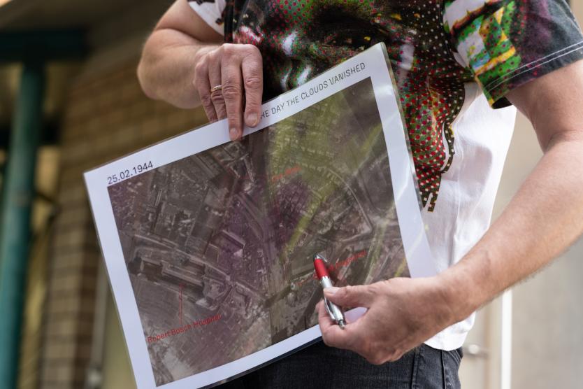 Nahaufnahme einer Person, die eine ausgedruckte Karte mit dem Datum 25.02.1944 und der Aufschrift „THE DAY THE CLOUDS VANISHED“ hält und mit einem Stift darauf zeigt.  Close-up of a person holding a printed map labeled "25.02.1944" and "THE DAY THE CLOUDS VANISHED," pointing at a detail with a metal pen.