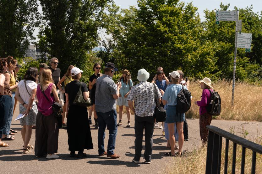 Eine Gruppe von Personen steht im Freien auf einem Weg bei Sonnenschein zusammen, umgeben von Bäumen und einem Wegweiser.  A group of people gathers outdoors on a sunny day on a paved path next to a green tree line and a signpost. 