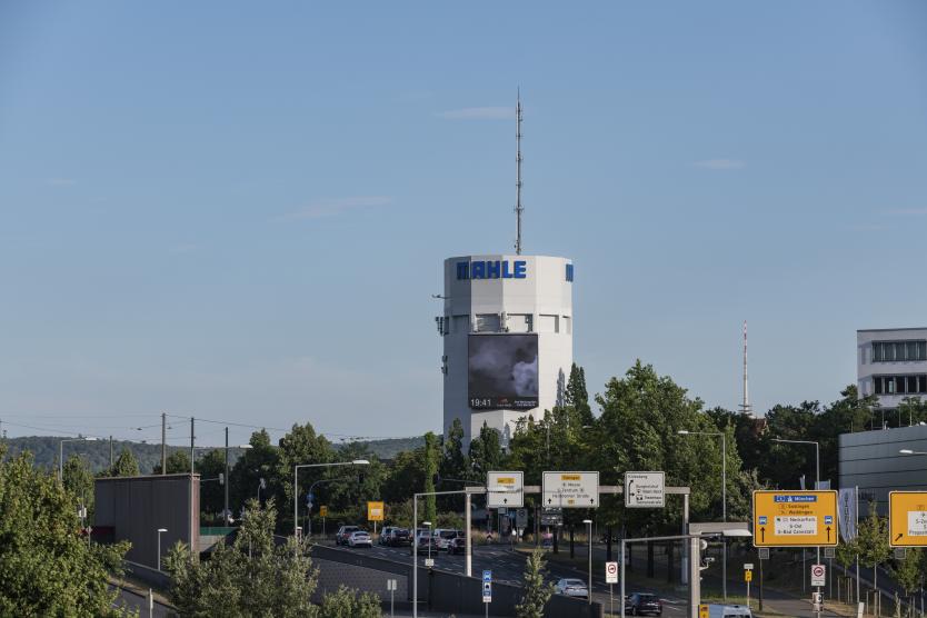 Panoramaansicht eines städtischen Gebiets mit einem hoch aufragenden Wasserturm der Firma MAHLE, an dem eine große digitale Werbefläche angebracht ist.  Wide view of an urban landscape featuring a tall MAHLE water tower fitted with a large digital billboard.