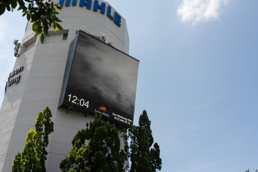Froschperspektive auf die massive Betonstruktur eines Turms mit der Aufschrift MAHLE und einem großen Bildschirm, der Wolkenmotive und die Uhrzeit 12:04 anzeigt.  Low-angle shot of a tall concrete tower displaying the MAHLE logo and a large screen showing a cloud image and the time 12:04.
