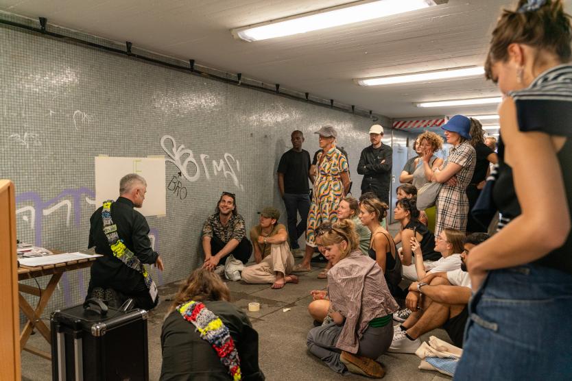 Eine Gruppe von Menschen sitzt auf dem Boden einer gefliesten Unterführung und nimmt an einer Veranstaltung oder Präsentation teil, während ein Mann an der Wand zeichnet.   A group of people sits on the floor of a tiled pedestrian underpass, attending an event or presentation while a man draws on the wall. 