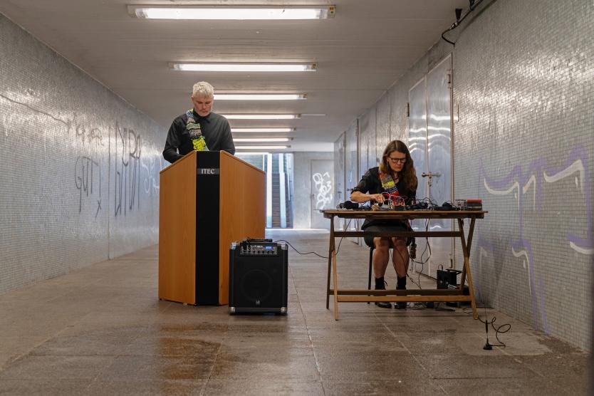 Zwei Personen stehen in einem gefliesten Tunnel; ein Mann spricht an einem hölzernen Rednerpult mit Lautsprecher, während eine Frau an einem Tisch mit technischen Geräten sitzt.   Two people stand in a tiled tunnel; a man speaks at a wooden lectern with a speaker, while a woman sits at a table with technical equipment.