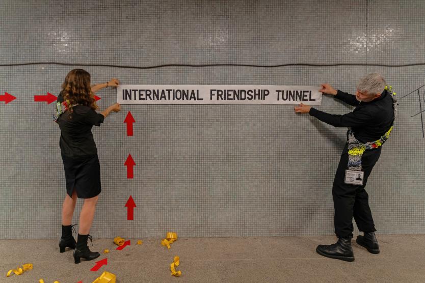 Zwei Personen bringen ein weißes Banner mit der Aufschrift „INTERNATIONAL FRIENDSHIP TUNNEL“ an einer gefliesten Wand in einem Tunnel an.   Two people attach a white banner reading "INTERNATIONAL FRIENDSHIP TUNNEL" to a tiled wall inside a tunnel.