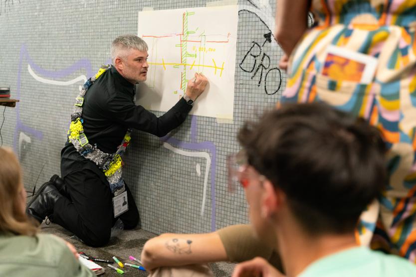 Ein Mann kniet vor einer gefliesten Tunnelwand, umgeben von Zuschauenden, und zeichnet mit einem Stift auf ein angeklebtes Papier mit einem bunten Diagramm. Er trägt eine auffällige, gewebte Textilschlinge über der Schulter.  A man kneels in front of a tiled tunnel wall, surrounded by spectators, drawing with a pen on a taped piece of paper featuring a colorful diagram. He wears a distinctive woven textile sling over his shoulder.