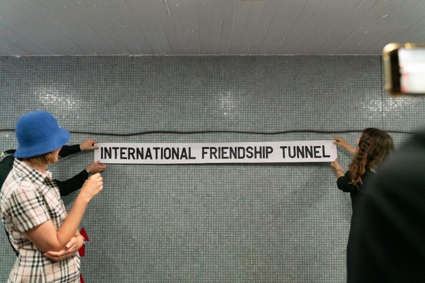 Zwei Personen befestigen ein langes weißes Schild mit der Aufschrift „INTERNATIONAL FRIENDSHIP TUNNEL“ an einer grauen, gefliesten Tunnelwand.  Two people attach a long white sign displaying the text "INTERNATIONAL FRIENDSHIP TUNNEL" onto a gray tiled tunnel wall. 