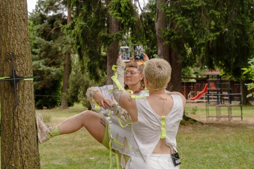 Zwei Performerinnen in hellen Outfits mit gelben Riemen stehen im Freien an einem Baum; eine stützt sich am Stamm ab, während die andere zwei Smartphones in die Höhe hält.  Two performers in light-colored outfits with yellow straps stand outdoors by a tree; one leans against the trunk while the other holds two smartphones up in the air.