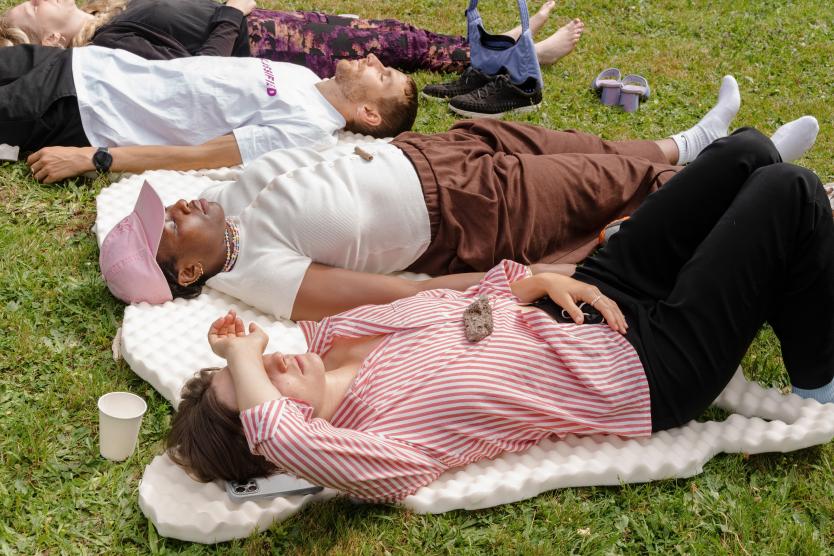 Mehrere Personen liegen entspannt auf Schaumstoffmatten auf einer Wiese im Freien und schauen nach oben.  Several people lie relaxed on foam mats in a grassy outdoor area, looking upward.