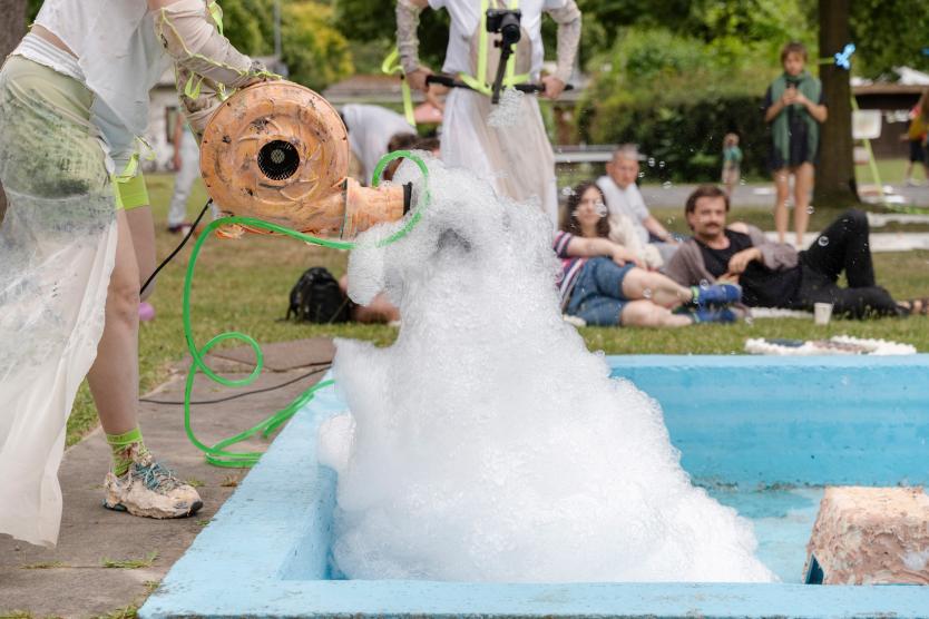 Ein oranges Gebläse füllt ein blaues Becken im Freien mit weißem Schaum, während Zuschauer im Hintergrund auf dem Rasen sitzen und zusehen.  An orange blower fills a blue outdoor basin with white foam, while spectators sit on the grass in the background watching the performance.